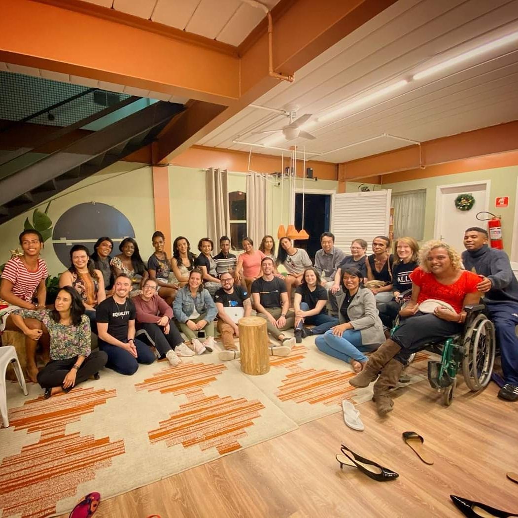 Grupo grande de mulheres acolhidas e equipe da Casa Florescer reunidos na sala de convivência, sentados no tapete e sorrindo para a foto.